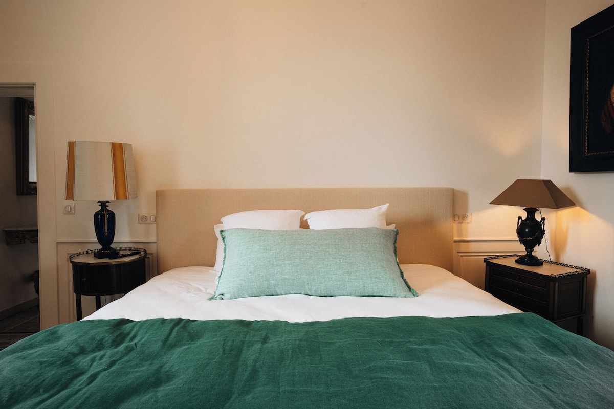 A neatly made bed with a green blanket and pillows, a bedside lamp, and a nightstand with a lamp.