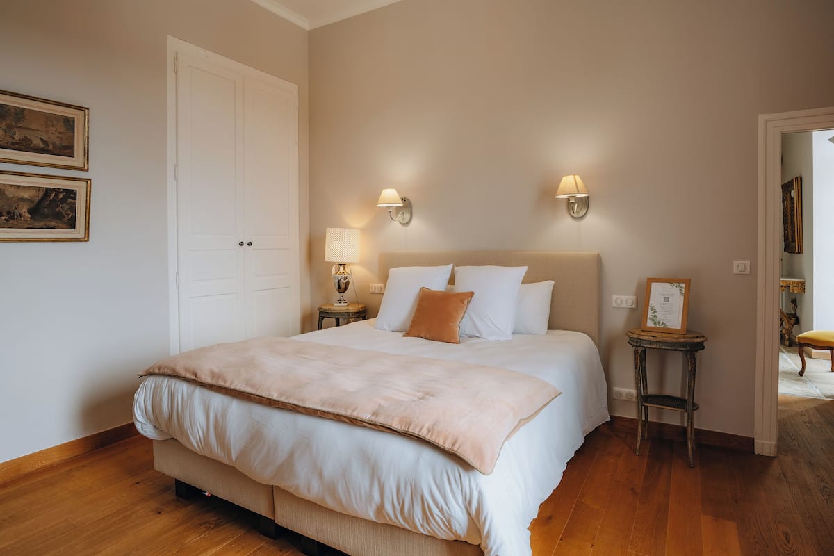 A bedroom with a large bed, two bedside tables with lamps, a wooden floor, and a door.