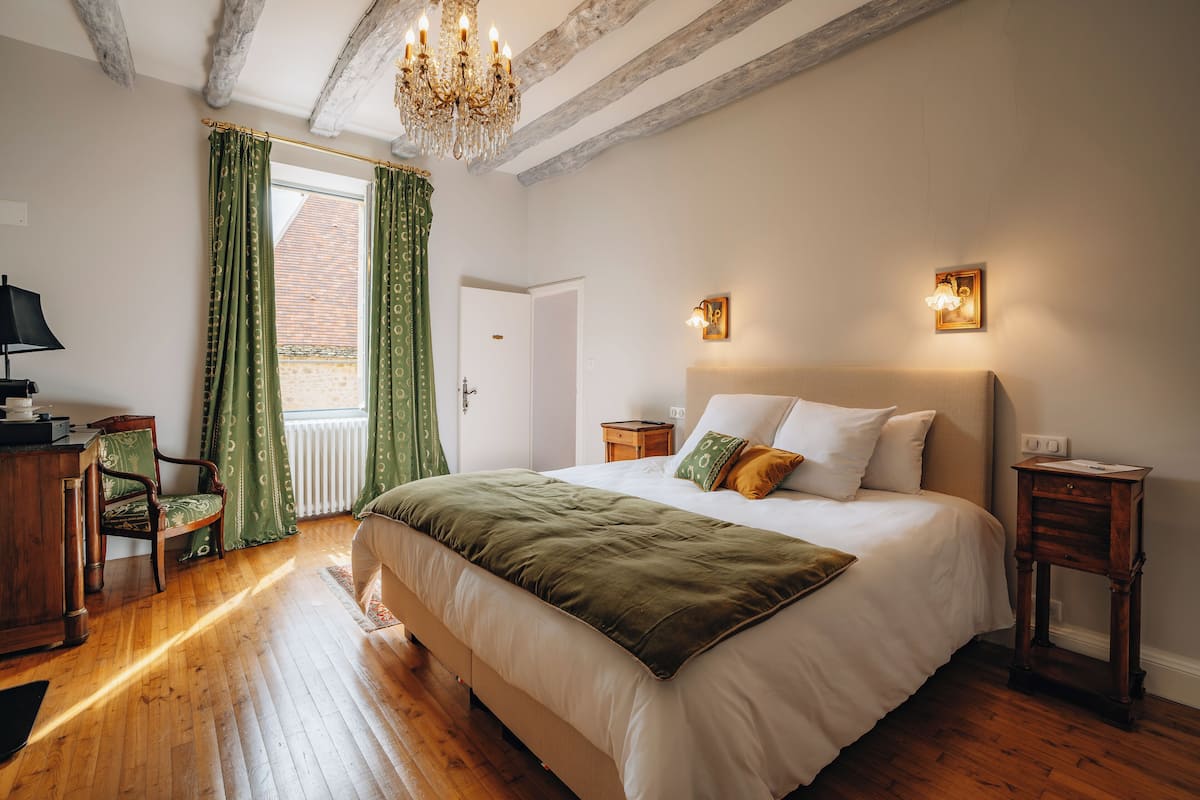 A bedroom with a large bed, wooden floor, green curtains, a chandelier, and a small table.