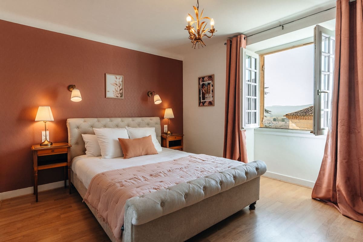 A bedroom with a large bed, two bedside tables with lamps, a chandelier, and a view through an open window.