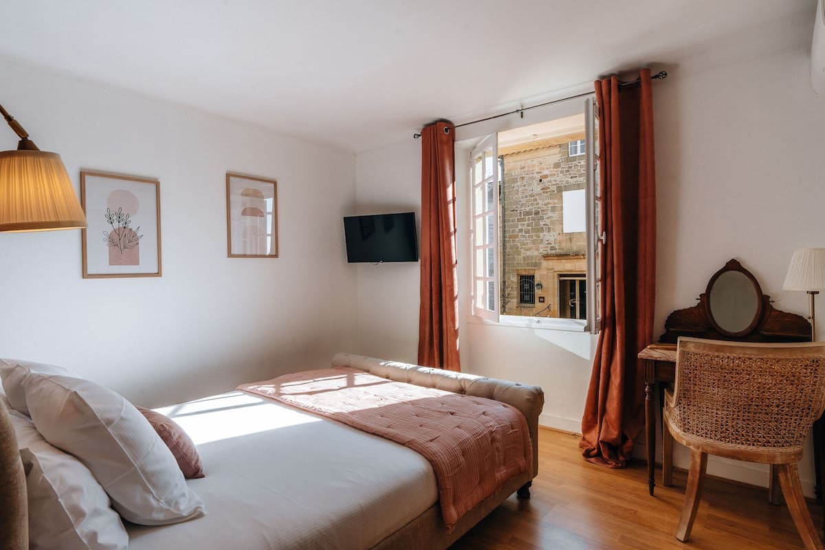 A bedroom with a bed, a desk with a mirror, a television, and a window with curtains.