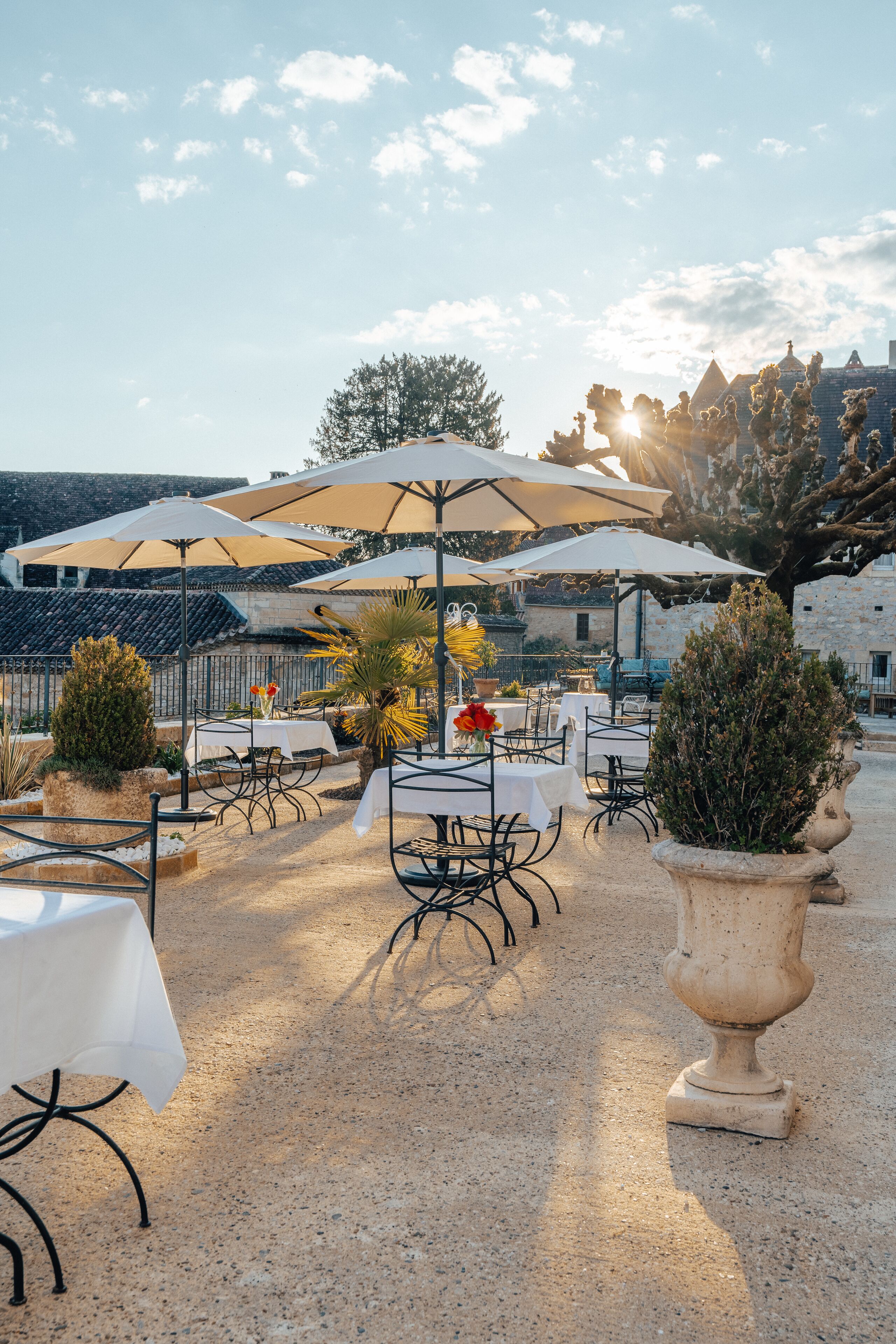An outdoor seating area with tables and umbrellas, surrounded by stone walls and potted plants.