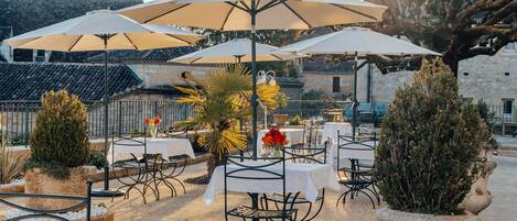 An outdoor seating area with tables and umbrellas, surrounded by stone walls and potted plants.