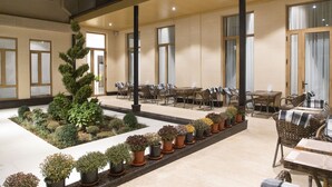 Um pátio interno espaçoso com diversos vasos de plantas, mobiliário de jardim com cadeiras e mesas de vime, e amplas janelas que proporcionam iluminação natural.