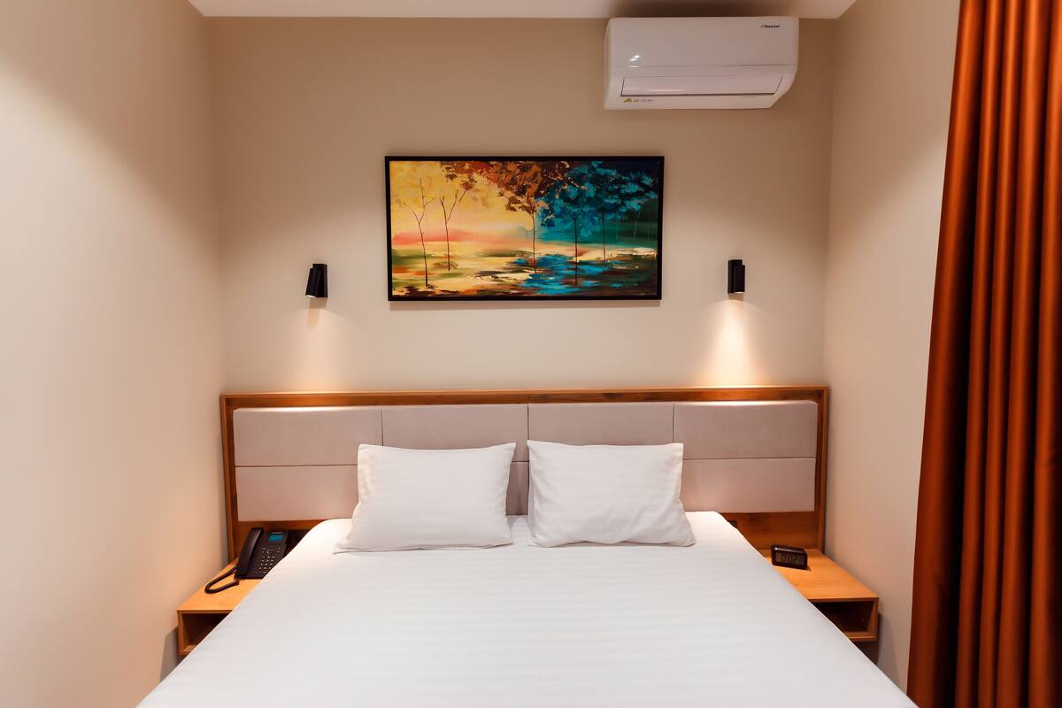 A hotel room with a bed, two bedside tables, a painting, an air conditioner, and a curtain.