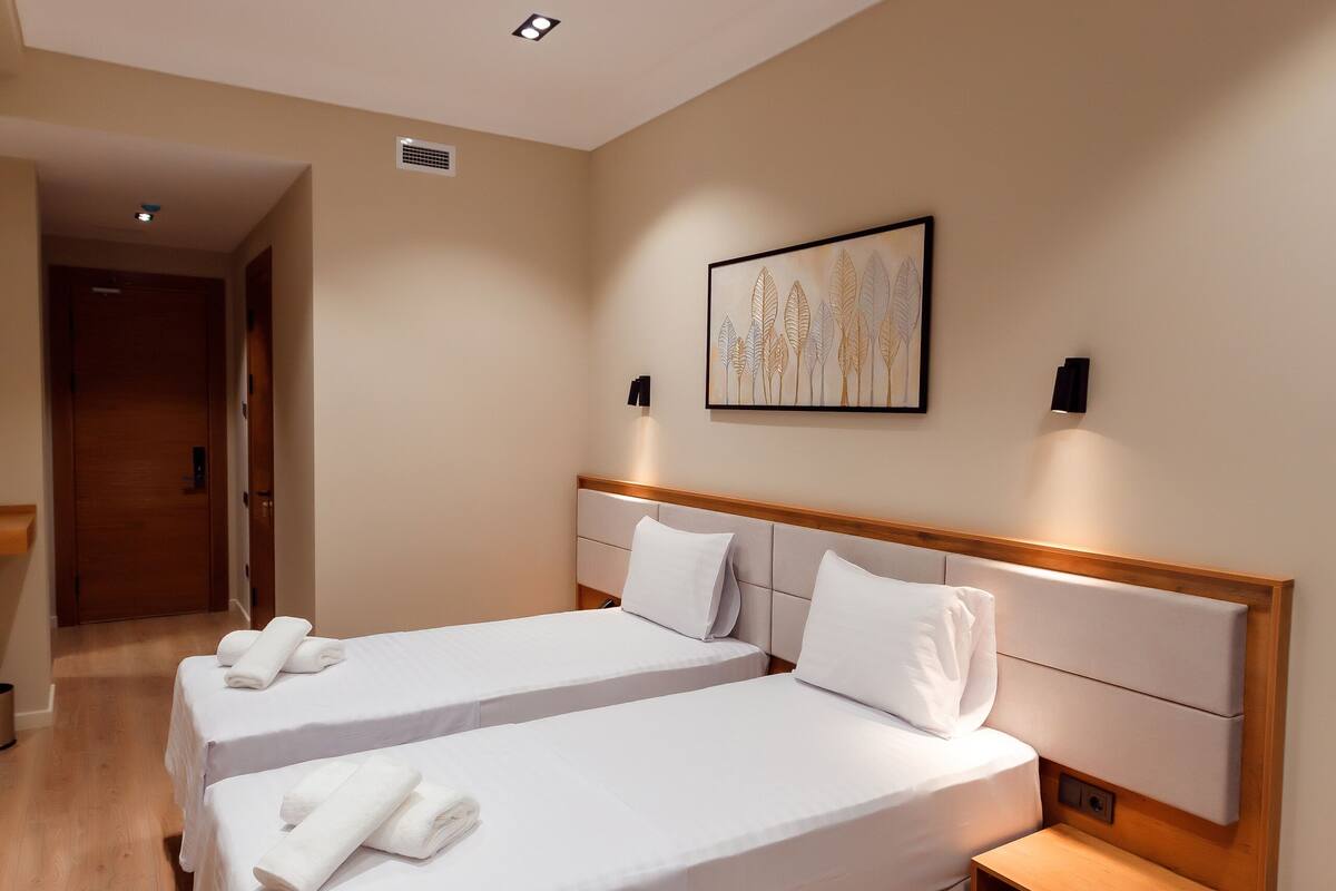 A hotel room with two beds, a wooden floor, a painting on the wall, and a nightstand with a lamp.