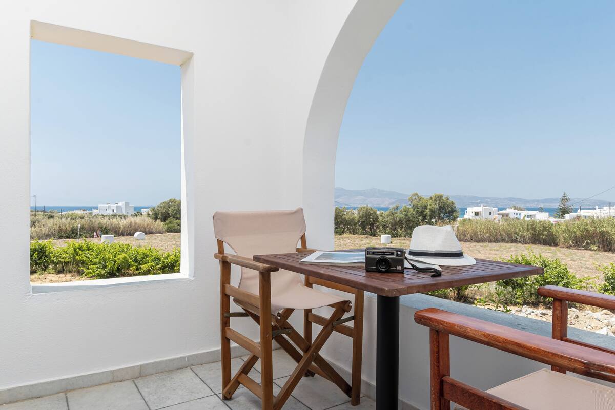 A balcony with a wooden table, a folding chair, and a view of the sea and buildings.