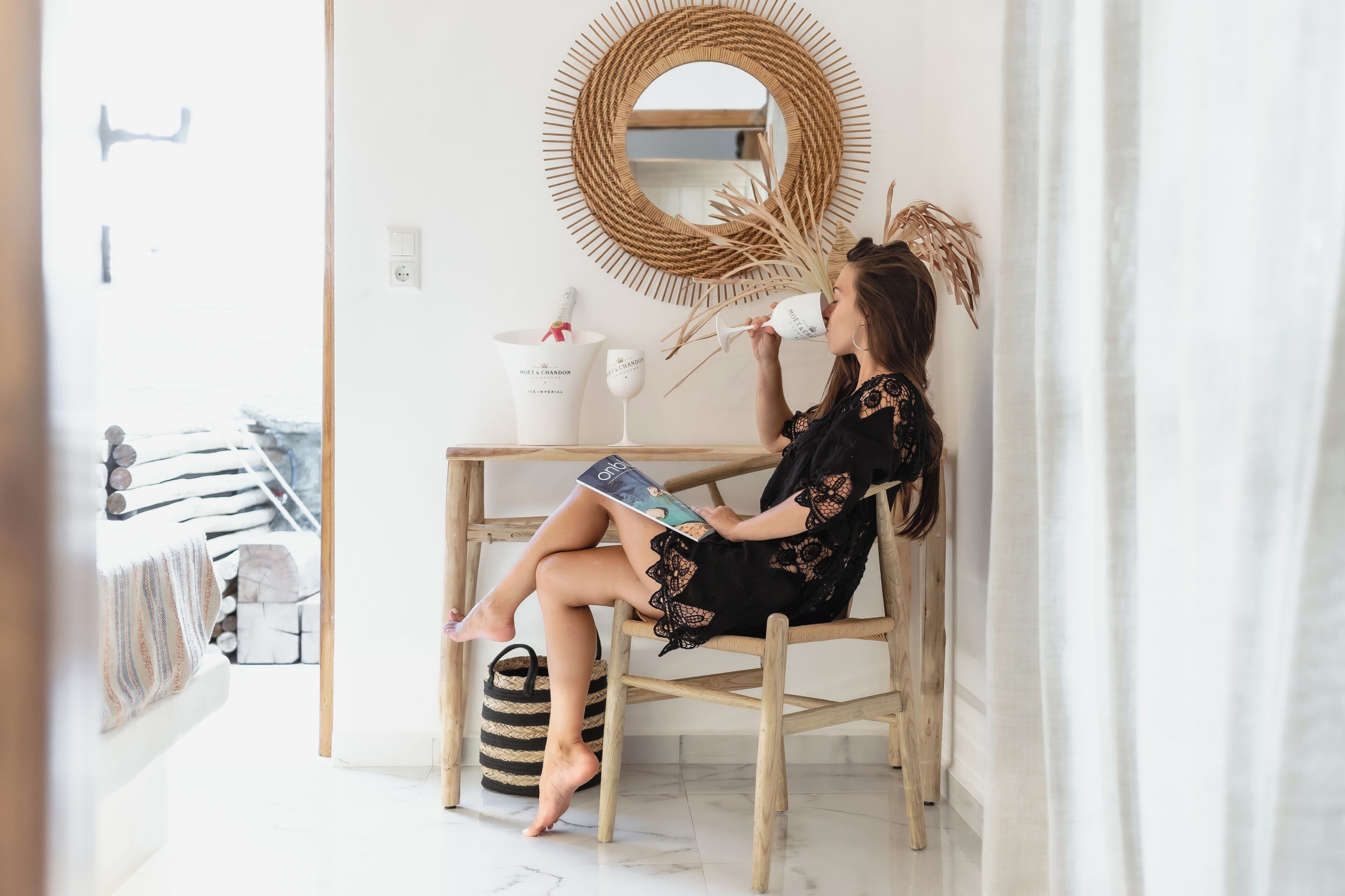 A woman sitting on a wooden chair in a room with a white wall, a round mirror, and a wicker basket.