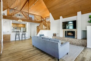 Un salón amplio con techo alto, vigas de madera, chimenea y una cocina al fondo.
