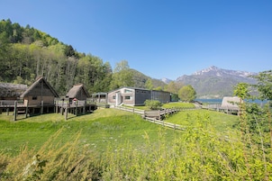 Una casa tradizionale in legno con tetto di paglia, un edificio moderno con ampie finestre e un pontile in legno che si affaccia su un lago con montagne sullo sfondo.