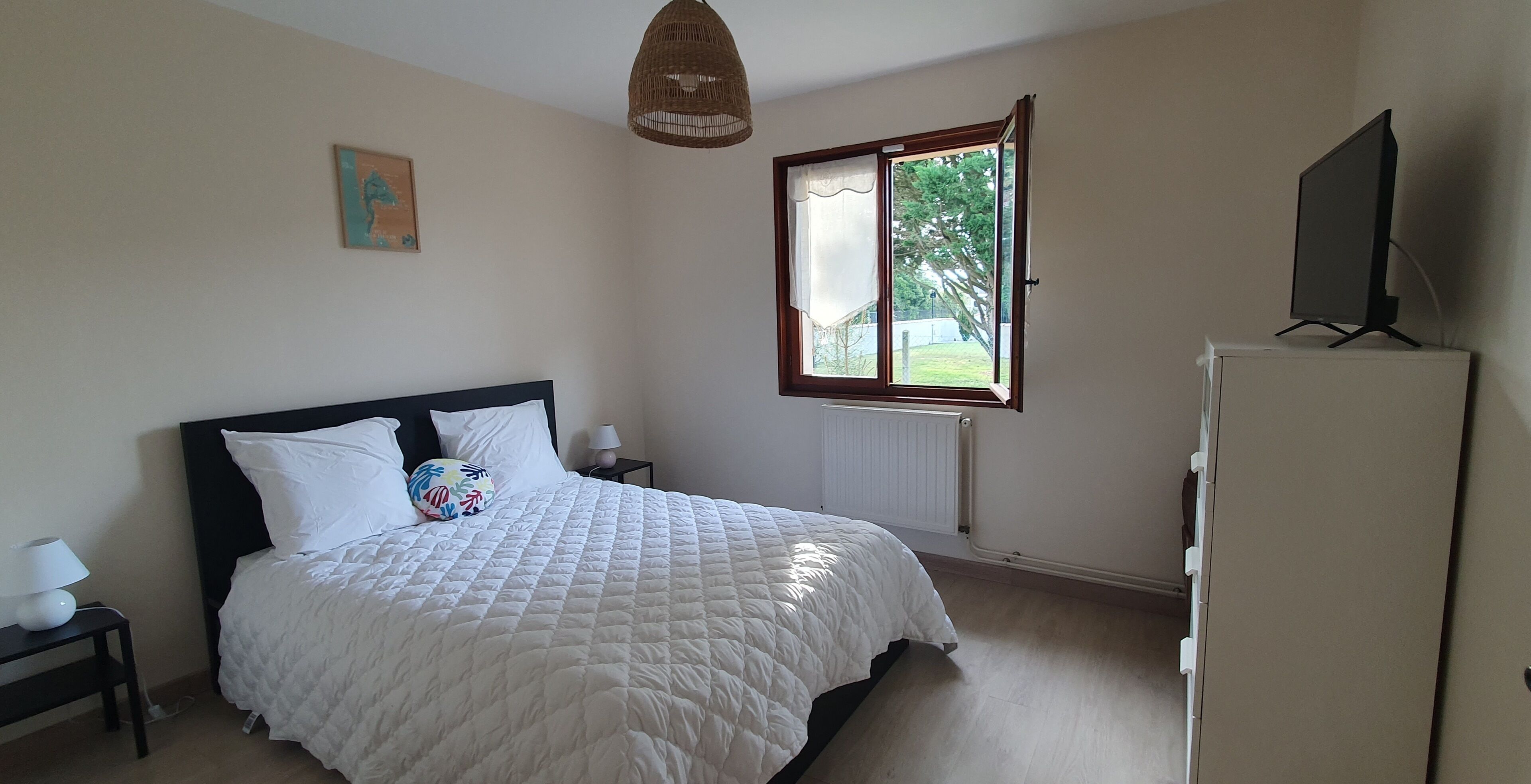 Ein Schlafzimmer mit einem Bett, einem Nachttisch mit Lampe, einem Fenster mit Vorhängen, einem Heizkörper und einem Fernseher auf einem Schrank.