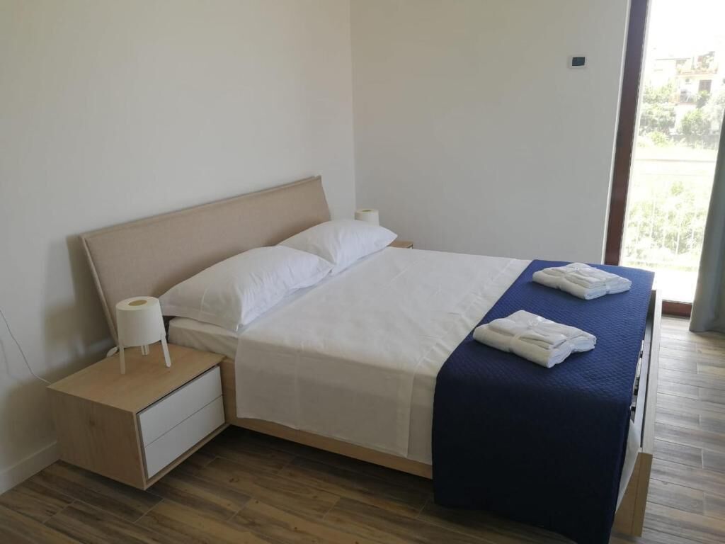 Un lit bien fait, avec du linge de lit blanc et une couverture bleue, une table de chevet en bois avec une lampe blanche, et une fenêtre donnant sur la verdure  à l’extérieur.