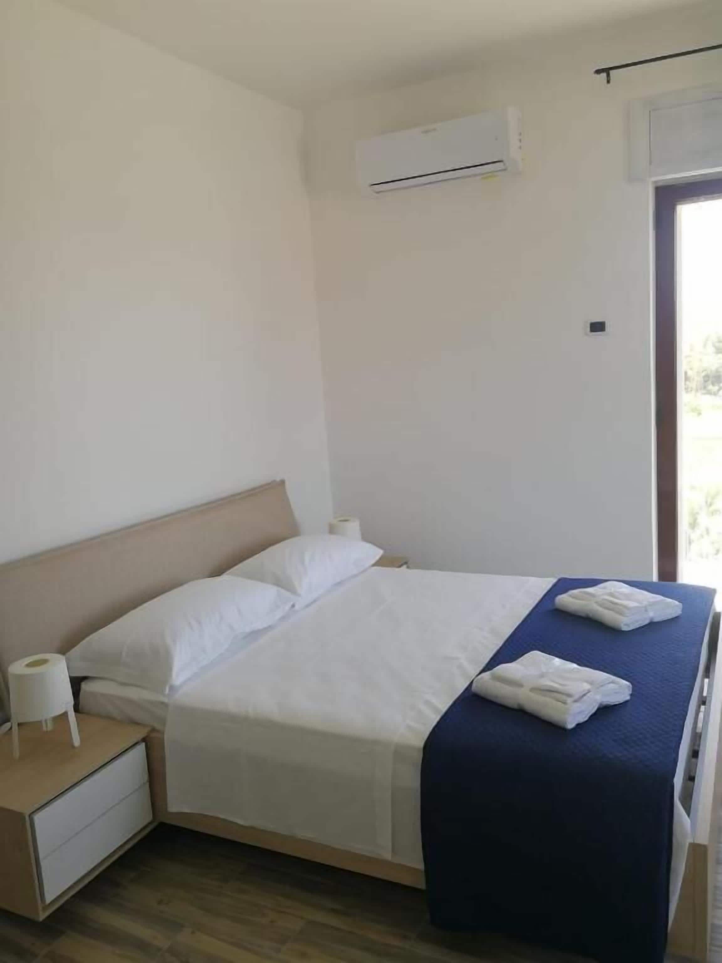 A neatly made bed with white linens, a blue blanket, and two towels on the side. There is a wooden nightstand with a white lamp and a roll of toilet paper beside the bed. An air conditioning unit is mounted on the wall above the bed.