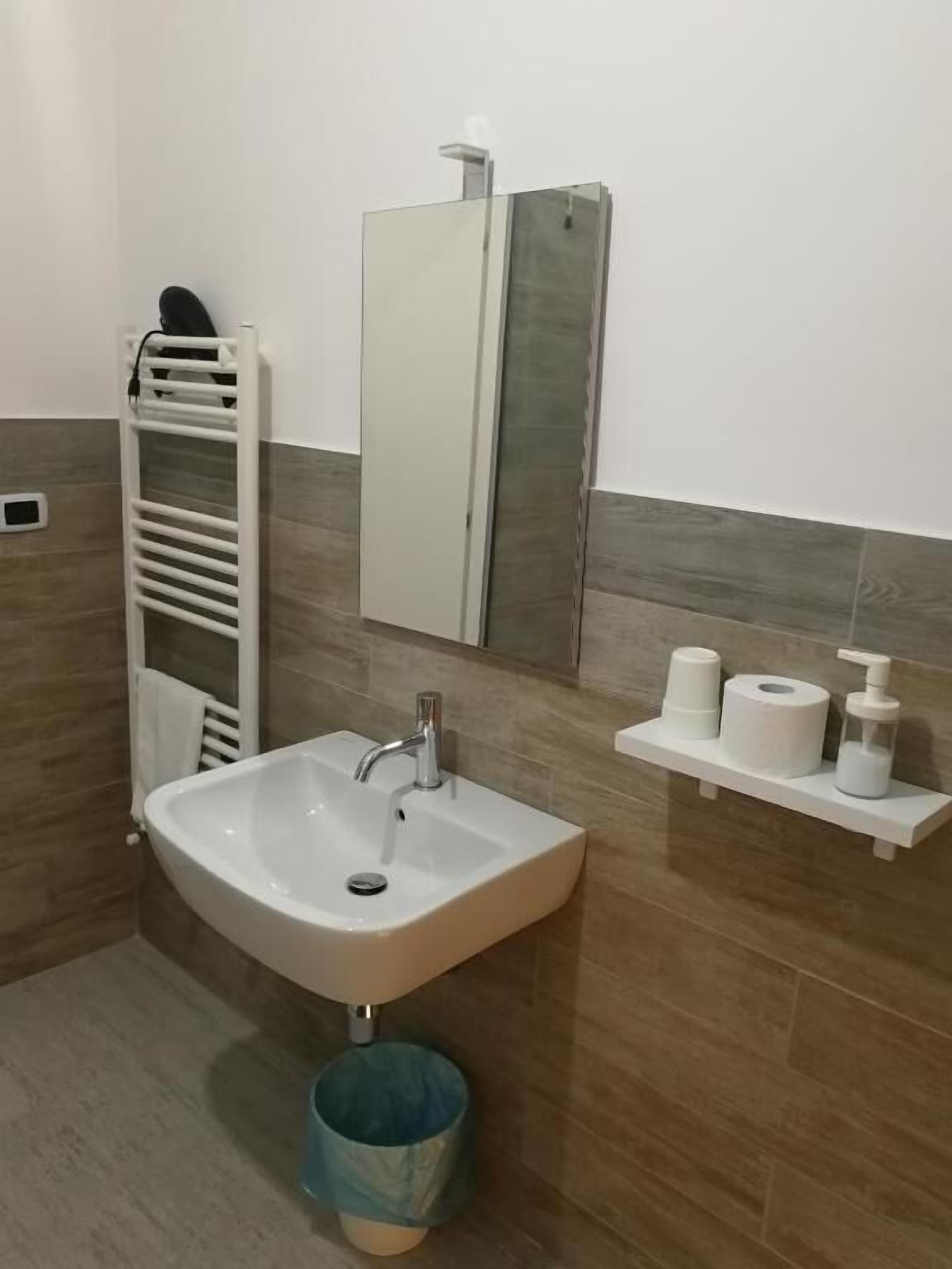 Un baño con lavamanos blanco, toallero, espejo y estante con artículos de aseo.