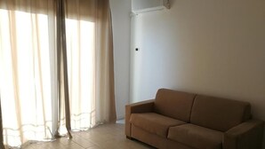 A living room with a beige sofa, a window with sheer curtains, and an air conditioning unit on the wall. - b&b Crispino (Belvedere Marittimo)