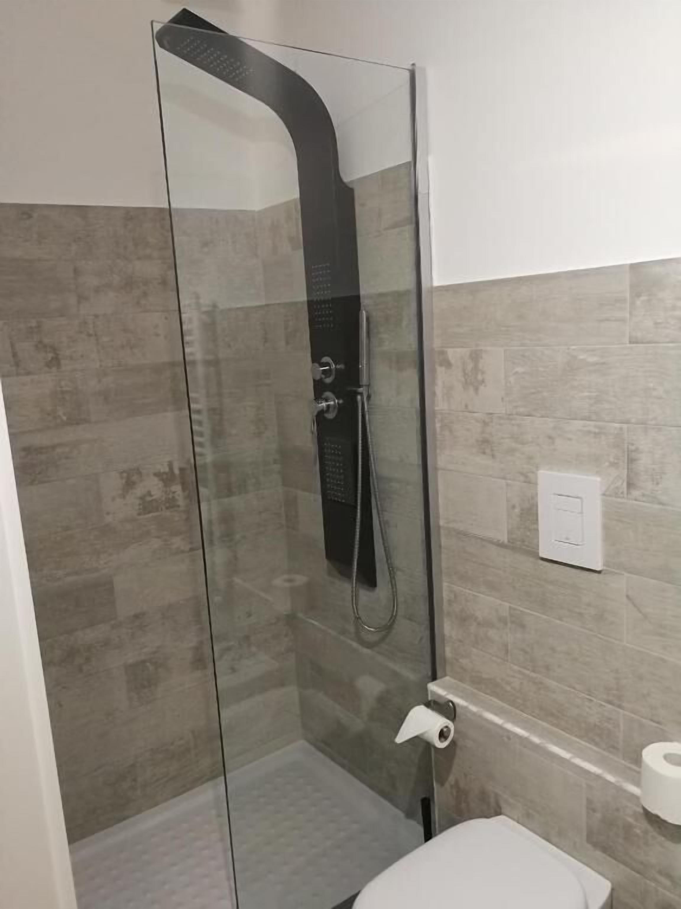 A glass-enclosed shower with a curved showerhead, a toilet, and a wall-mounted light switch.