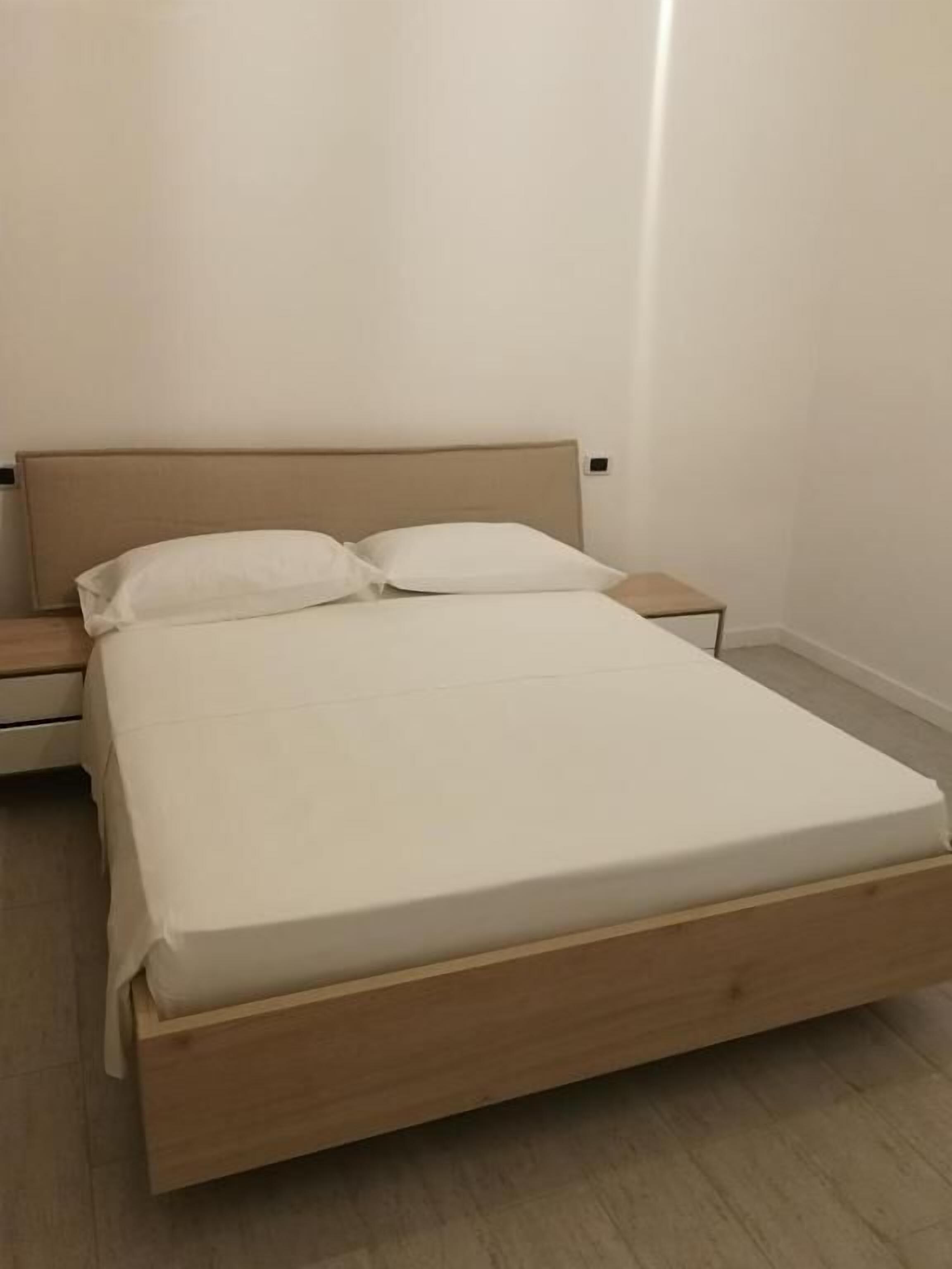 Una cama individual con estructura de madera y ropa de cama blanca en una habitación con paredes blancas lisas y suelo de madera.