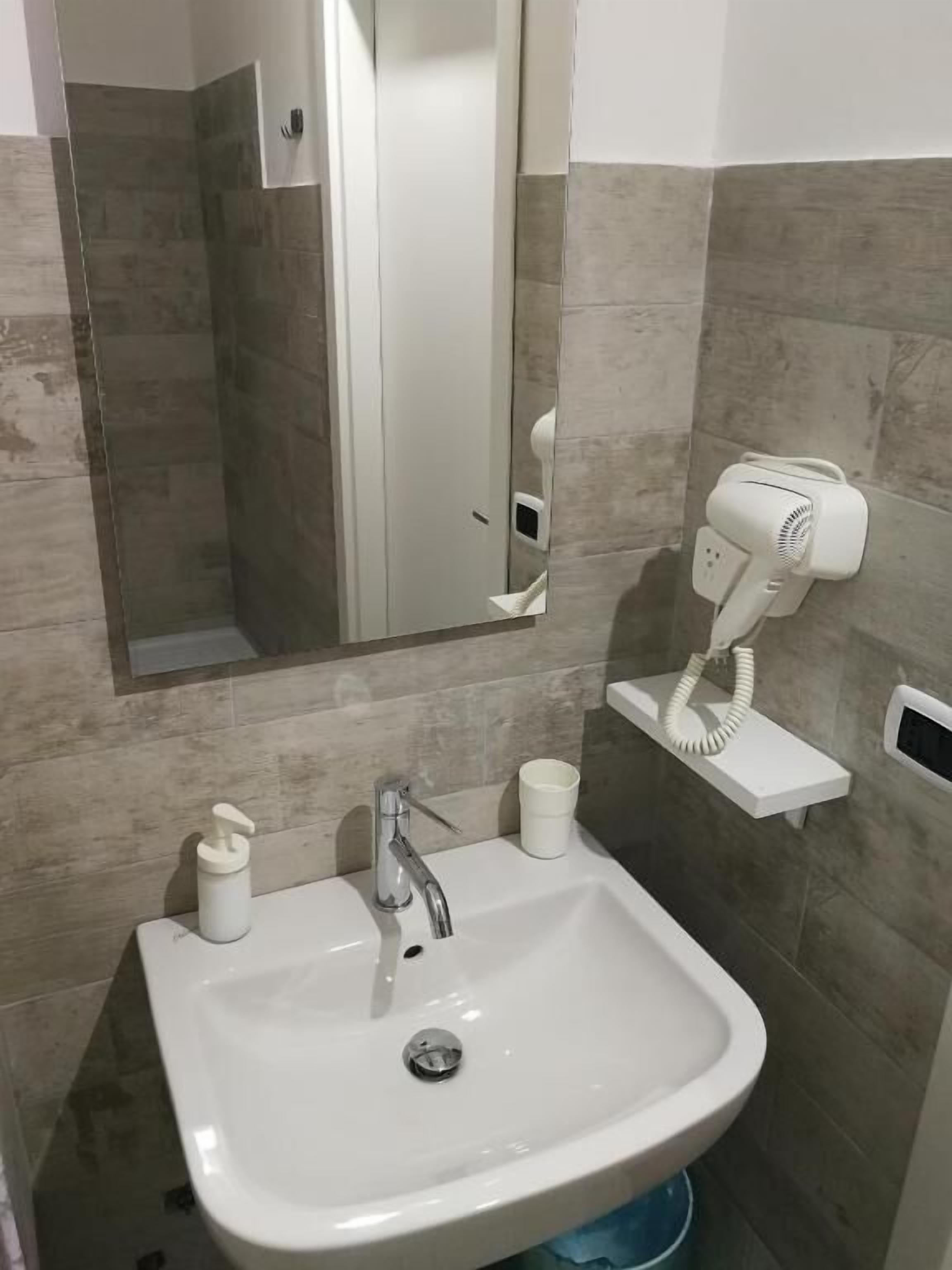 A bathroom with a white sink, a mirror, a hairdryer on a wall-mounted stand, and a small shelf with a cup.