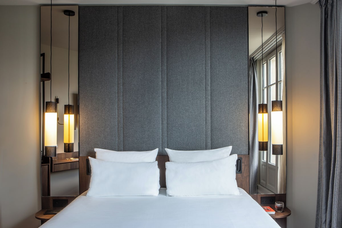 A modern hotel room with a large bed, two bedside tables, a mirror, and a window with curtains.
