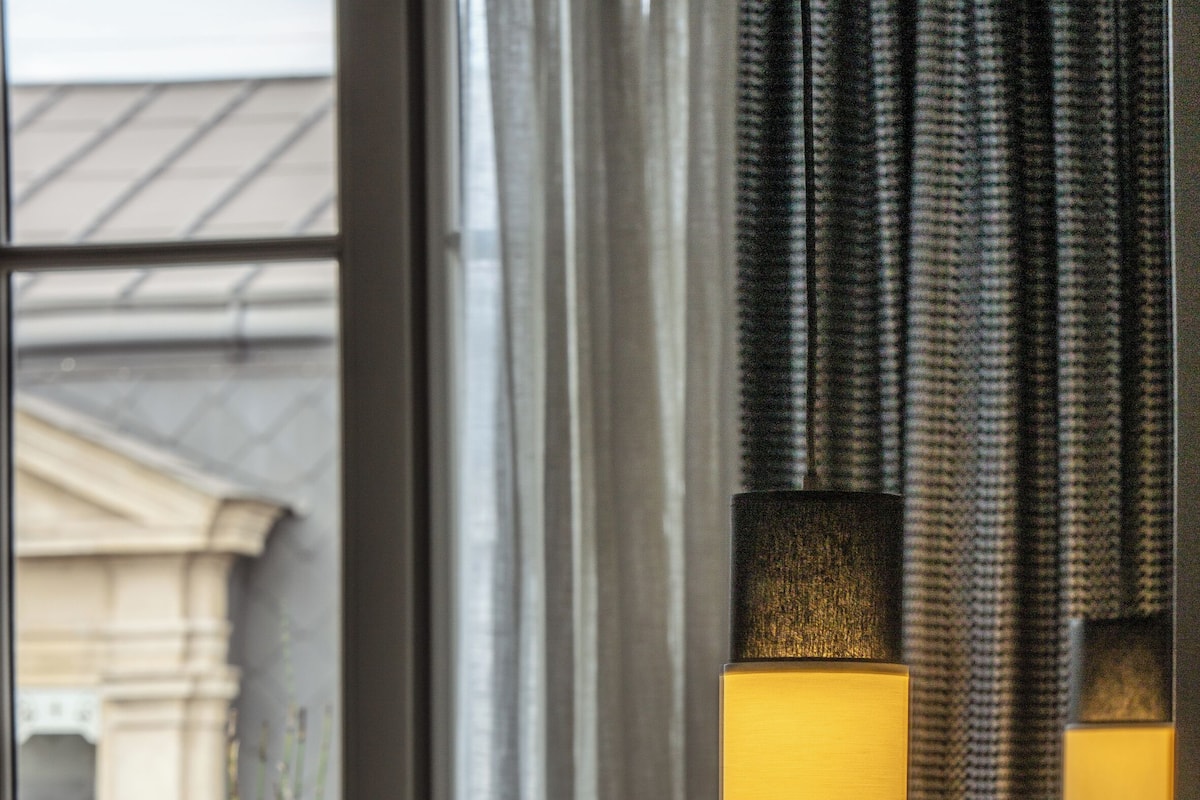 A room with a view of a building through a window, a lamp, and a cushion on a bed.