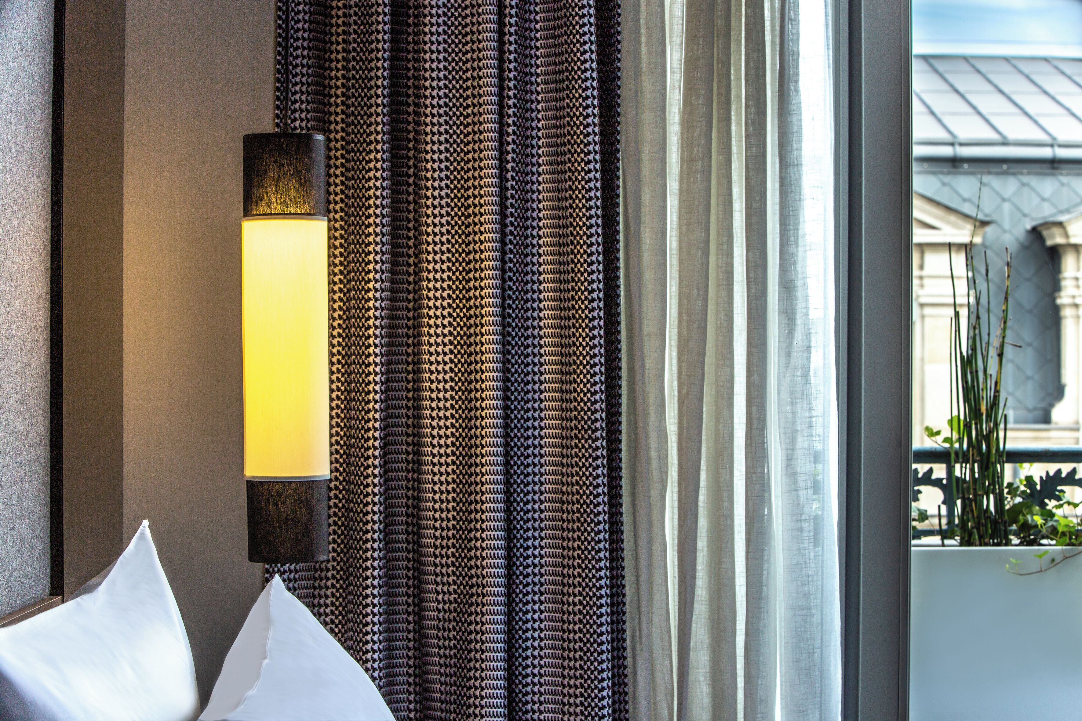 A hotel room with a bed, a wall-mounted lamp, patterned curtains, and a view of a building through a window.