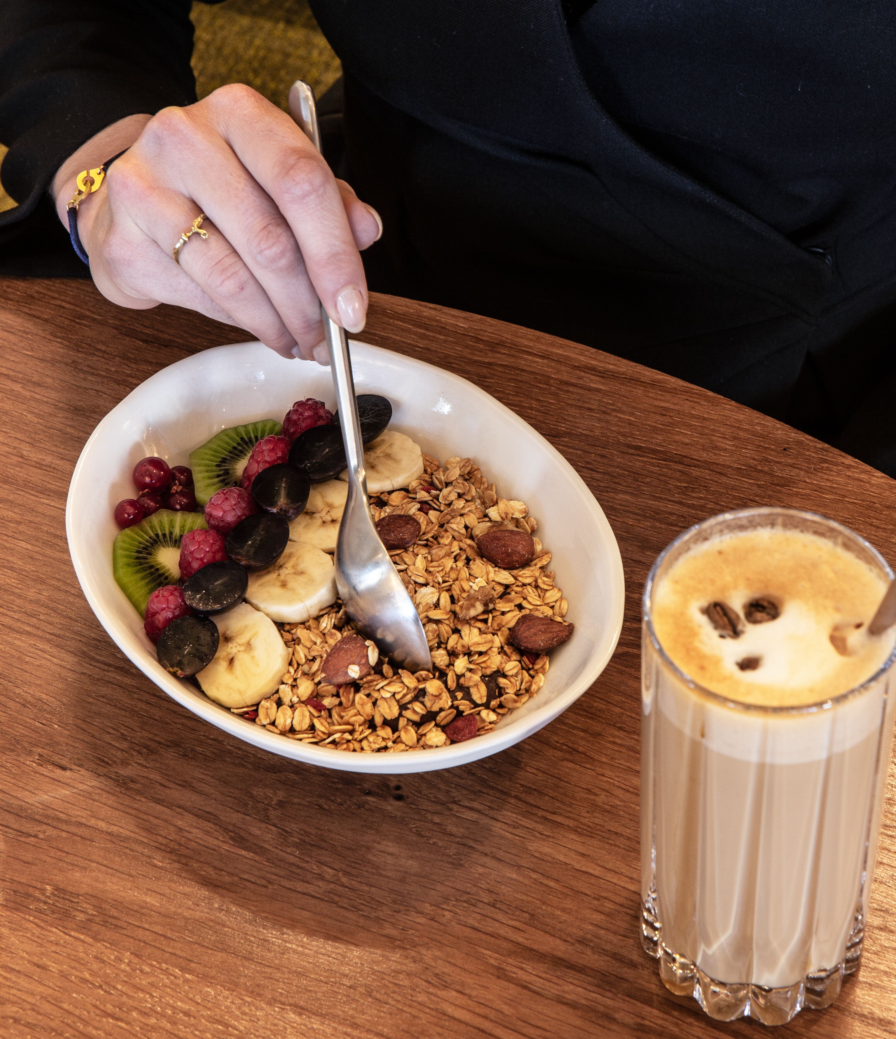 A bowl of granola with fruits and nuts, a spoon, and a glass of coffee on a wooden table.