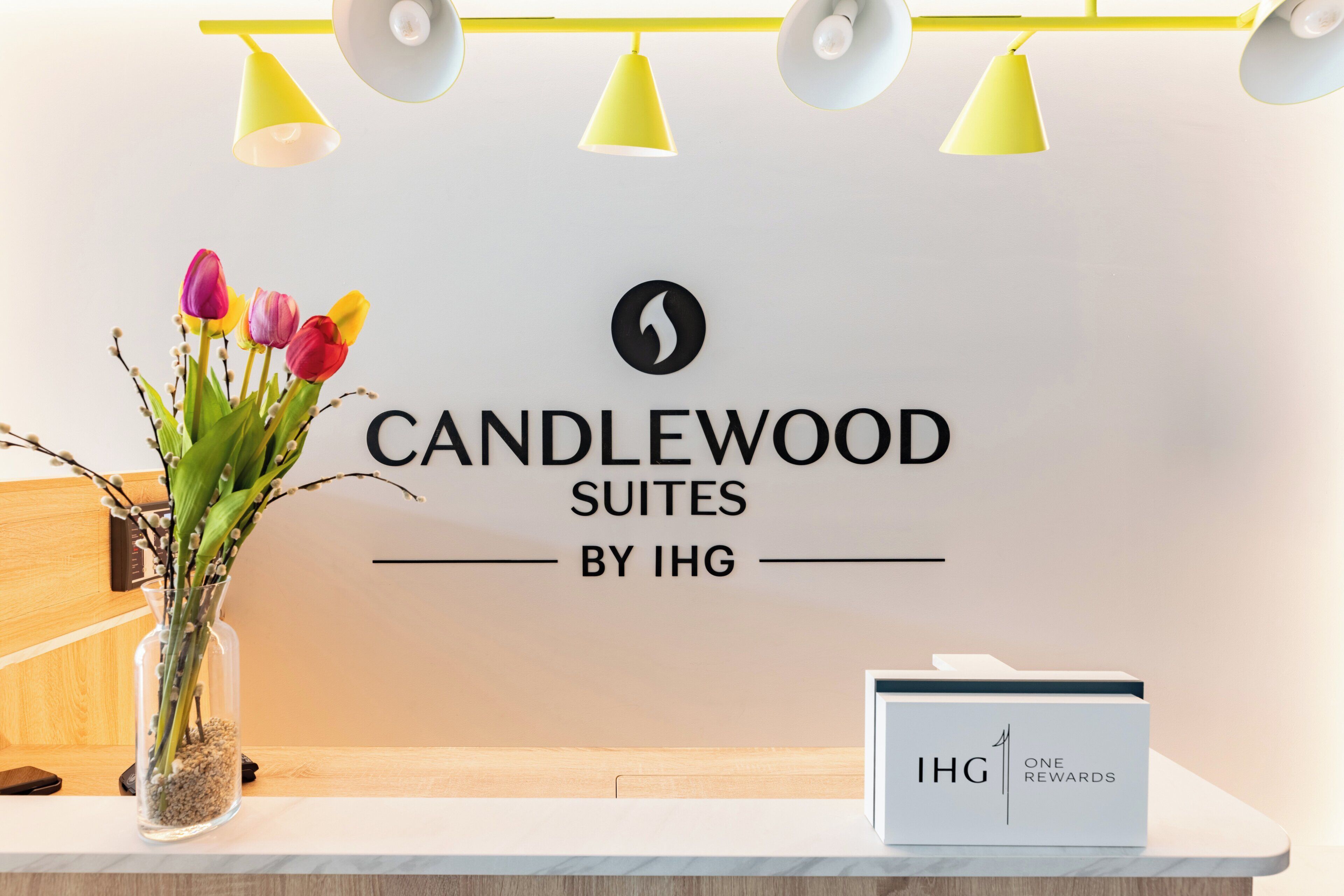 Una mesa de recepción con un florero, un letrero de Candlewood Suites by IHG y una caja que dice IHG One Rewards.