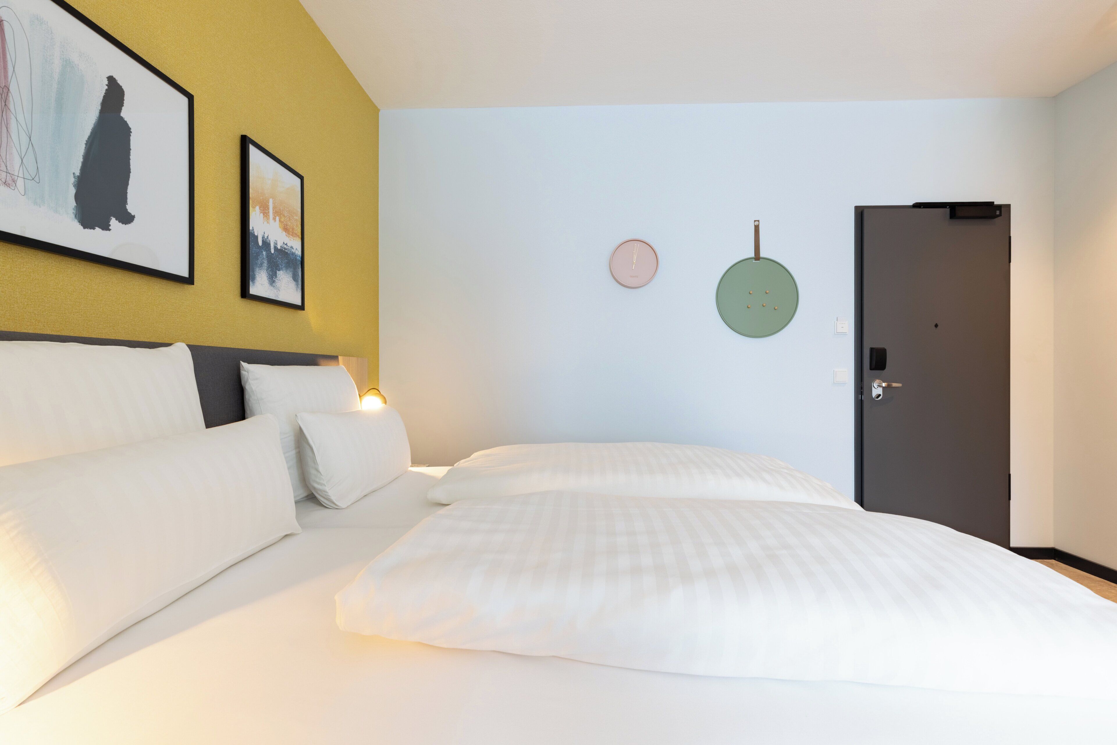 Quarto de hotel com uma cama, mesa de cabeceira, relógio de parede e uma porta.