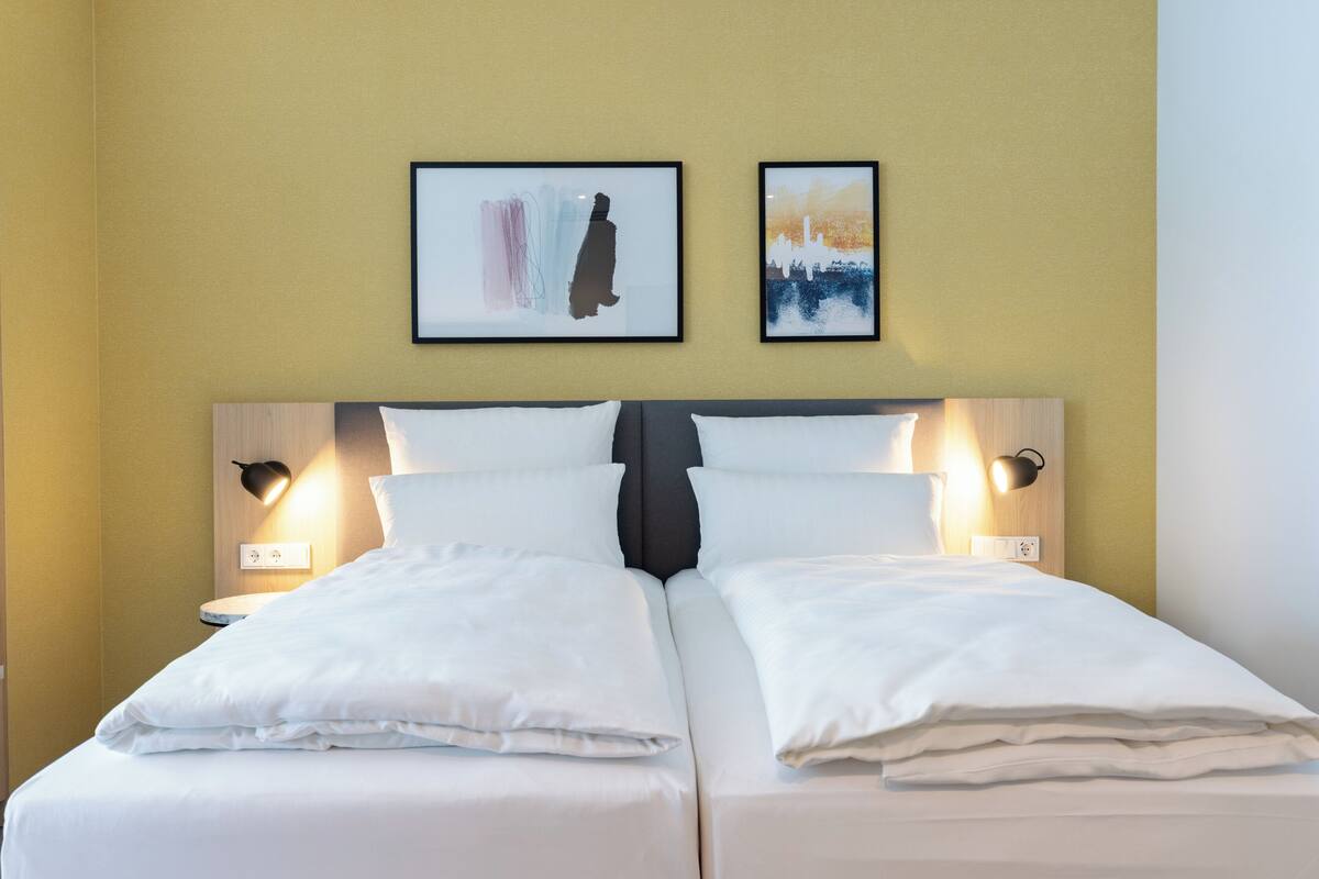 A double bed with white linens, two wall-mounted nightstands with lamps, and two framed artworks above the bed.