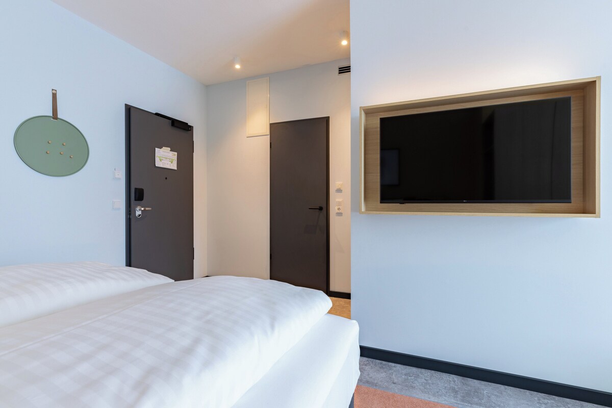 A hotel room with a bed, a door, a wall-mounted TV, and a light switch.
