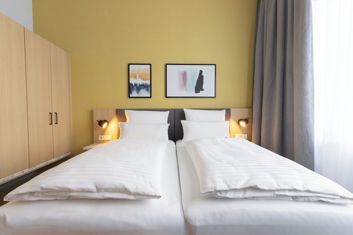 A double bed with white linens, two bedside tables with lamps, a wooden wardrobe, and two framed artworks on the wall.