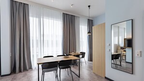 A modern hotel room with a dining area, large windows with curtains, a wooden door, and a mirror on the wall.