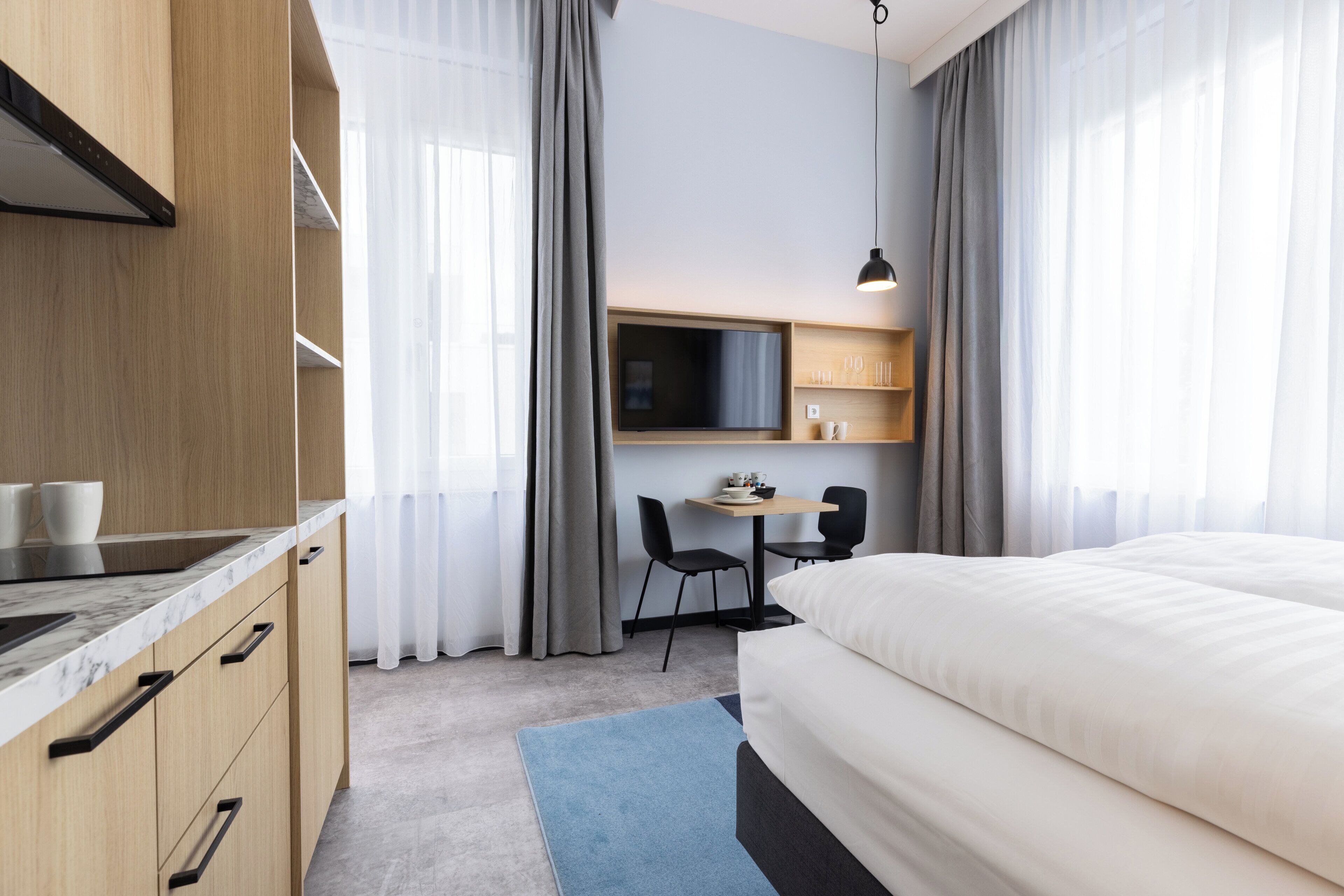 Habitación de hotel con cama, escritorio con sillas, televisor y zona de cocina con armarios y encimera.