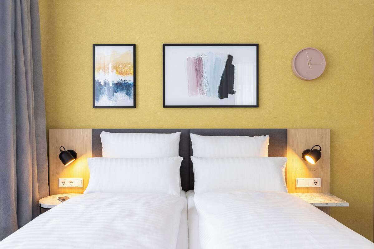 A double bed with white linens, two wall-mounted nightstands, two framed abstract artworks, a round wall clock, and a bed with a black headboard.