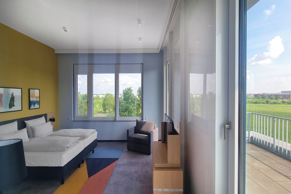 A modern bedroom with a large bed, a chair, a TV, and a balcony with a view.