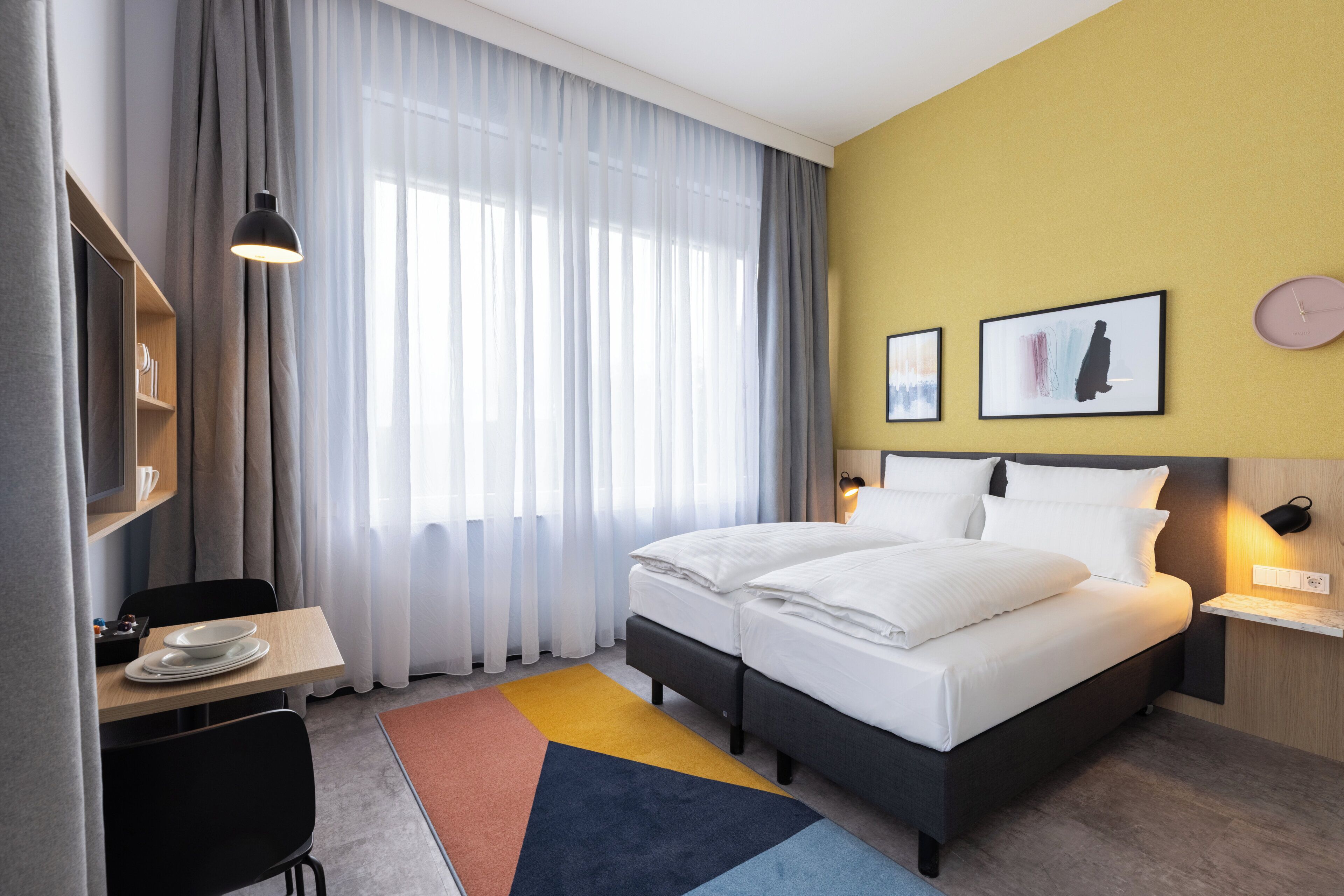 A hotel room with a bed, a desk with a chair, a small table with plates, a colorful rug, and a large window with curtains.