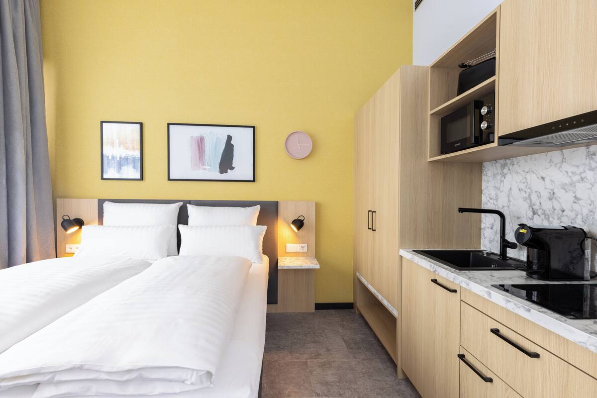 A hotel room with a bed, bedside tables, a small kitchenette, and two framed pictures on the wall.