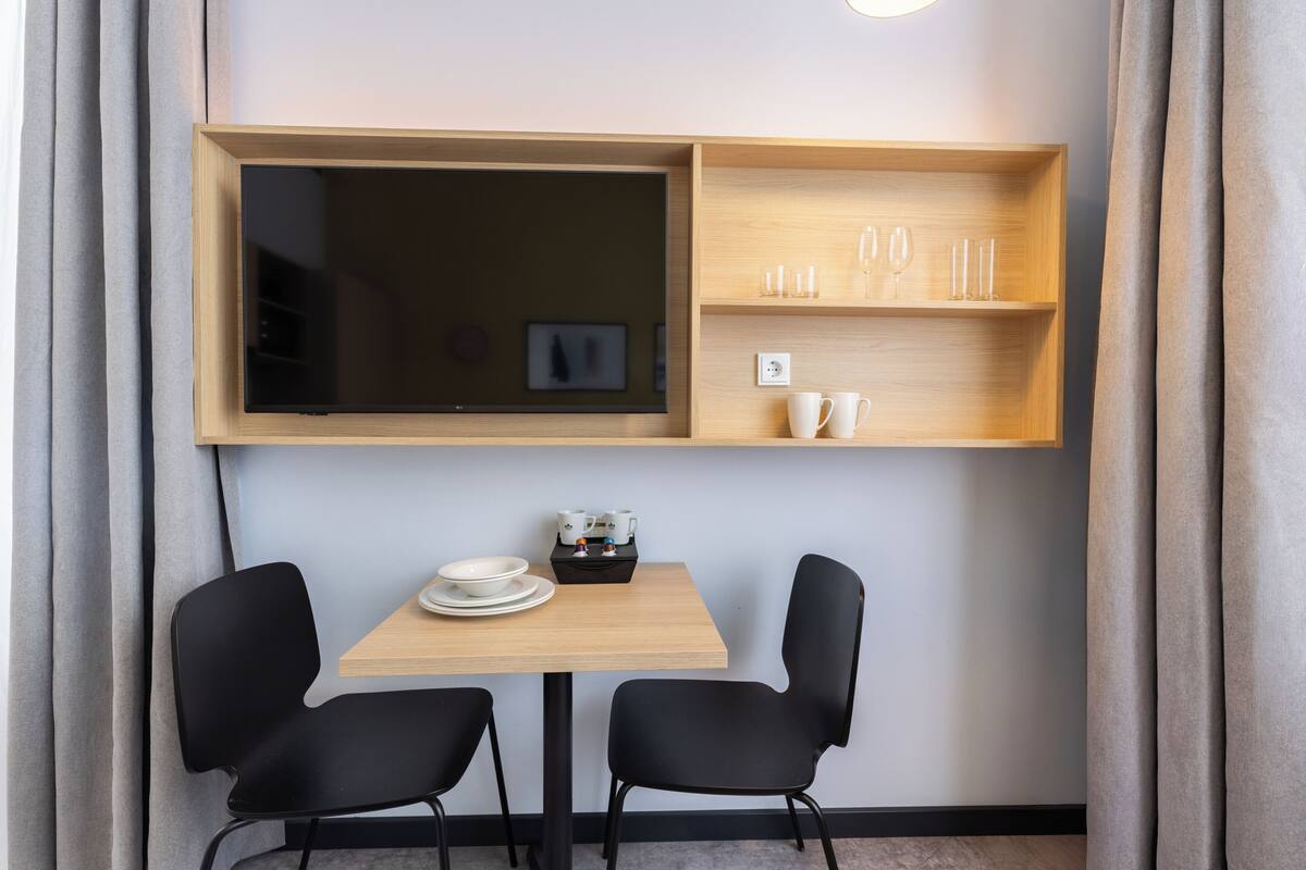 A small dining area with a wooden table, two black chairs, a shelf with glasses and cups, and a TV mounted above the table.