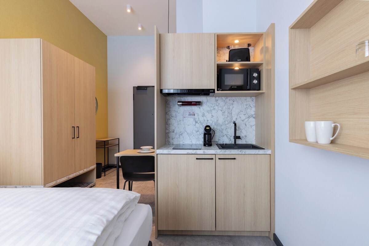 A compact hotel room with a kitchenette, a bed, a desk, and a chair.
