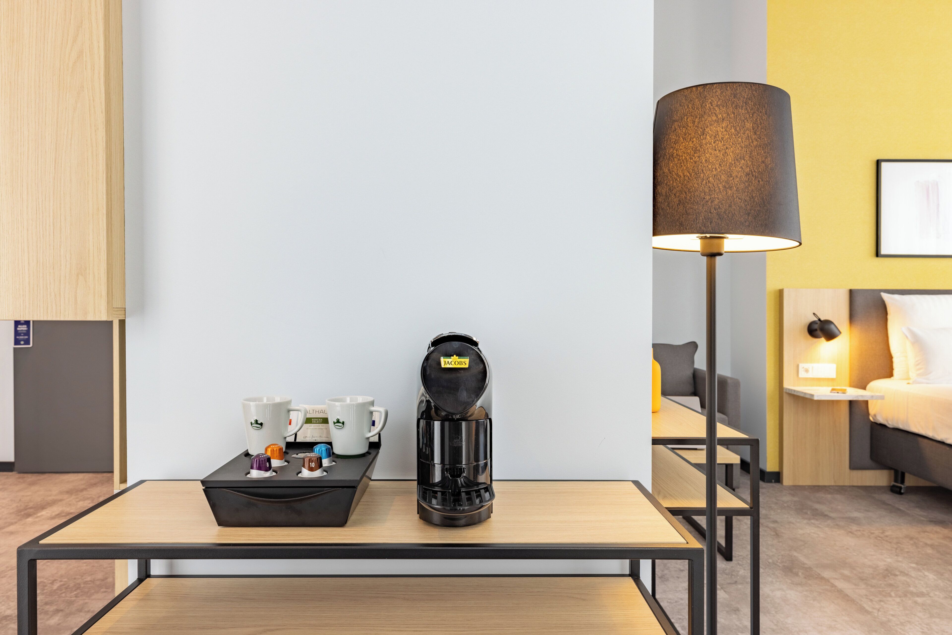 A hotel room with a coffee maker, a lamp, a bed, and a bedside table.