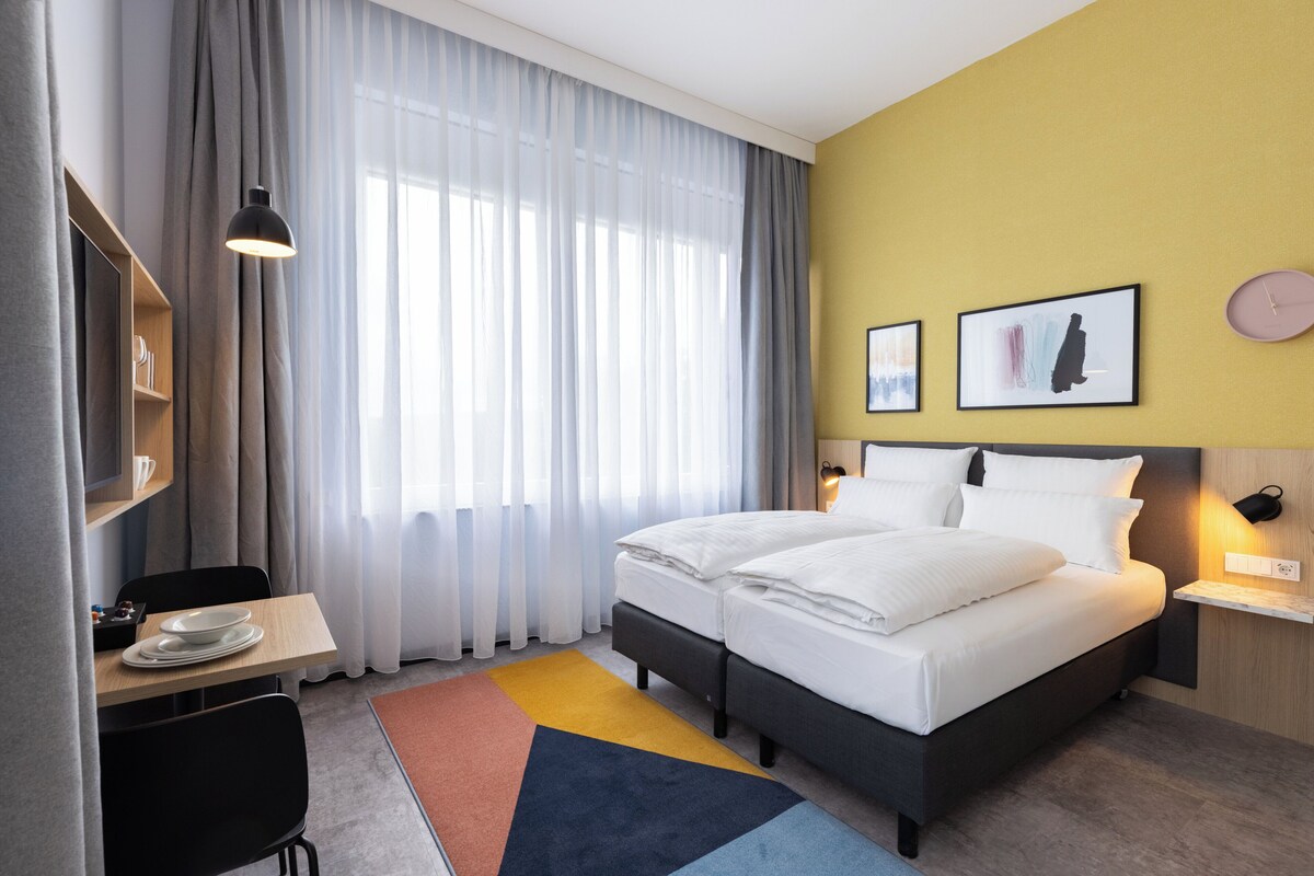 A hotel room with a bed, a desk with plates, a chair, a lamp, and a window with curtains.