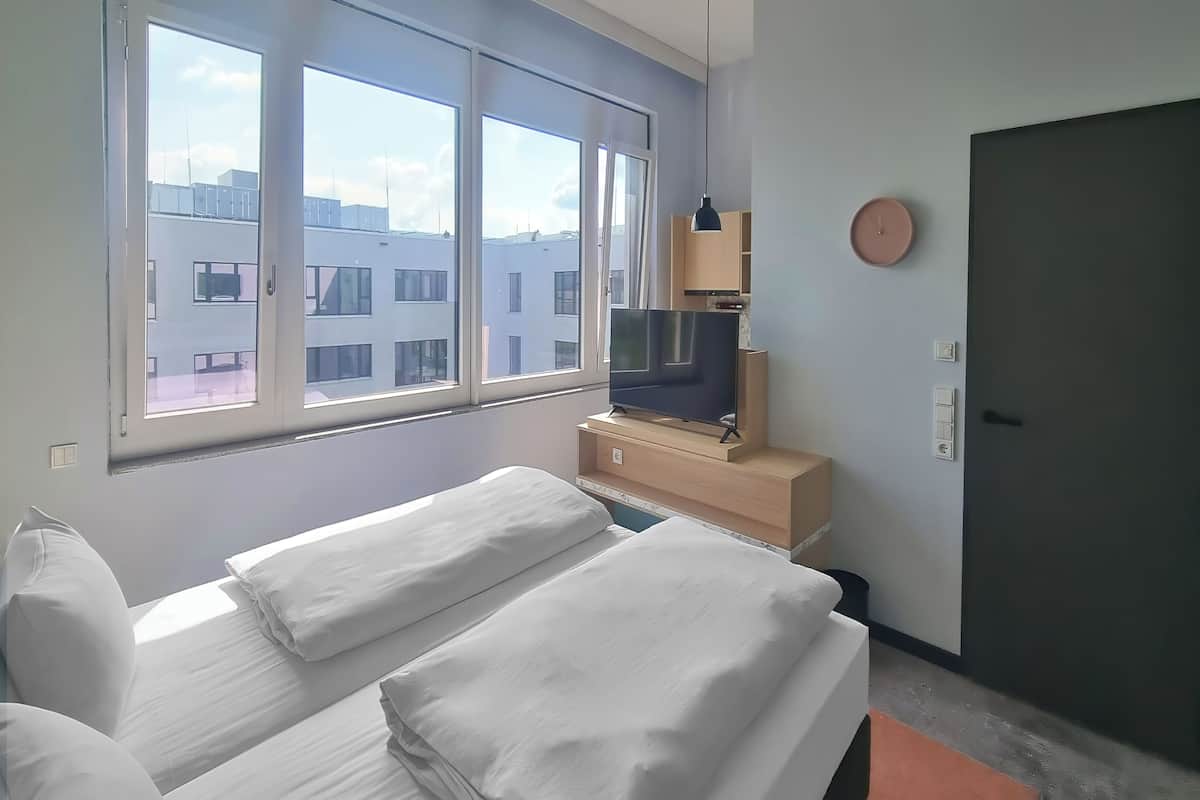Ein Hotelzimmer mit Bett, Fernseher auf einem Holzboden, Fenster mit Blick auf die Stadt und einer Tür.