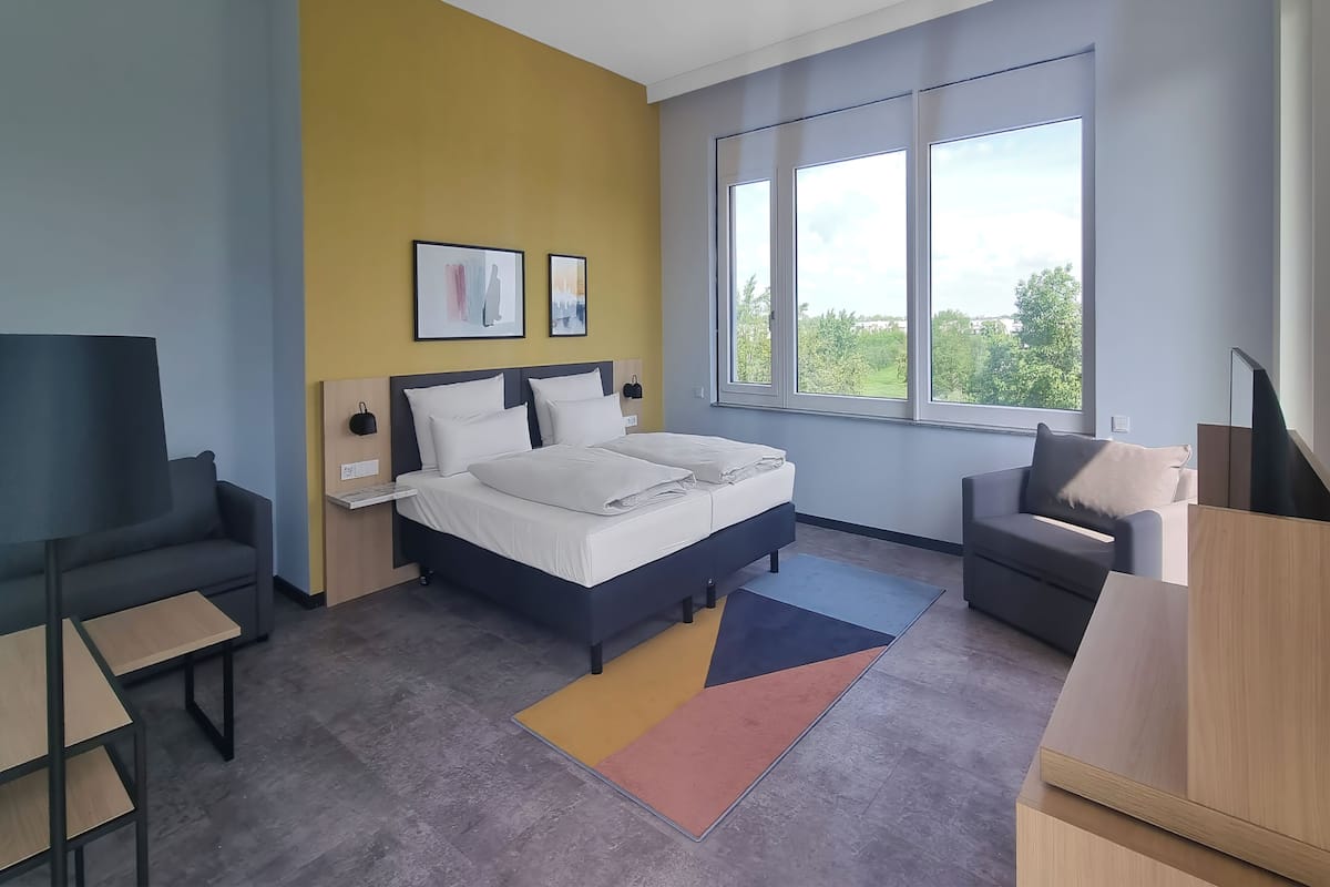 Ett modernt hotellrum med en säng, två soffor, en TV och stora fönster.