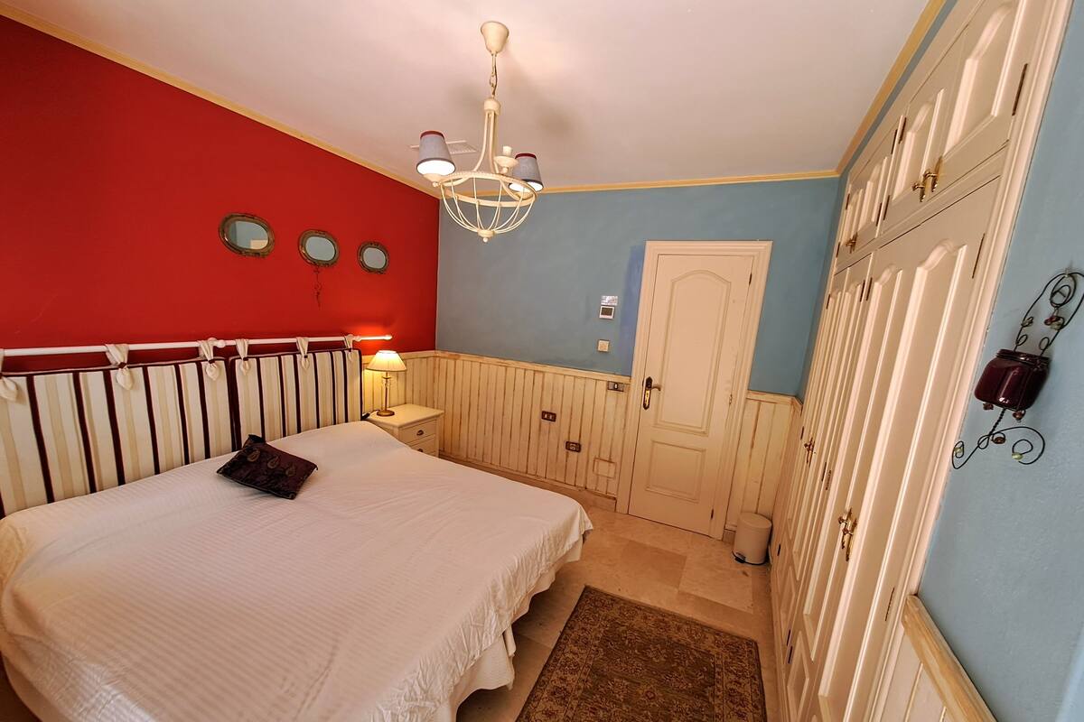 A bedroom with a bed, a nightstand, a chandelier, a door, and a wall-mounted light fixture.