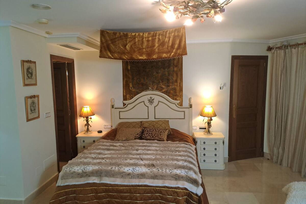 A bedroom with a large bed, two bedside tables with lamps, a chandelier, and a window with curtains.