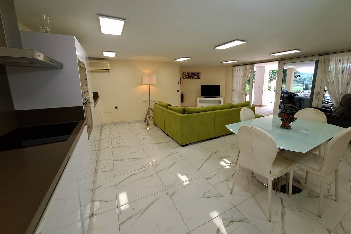 A modern living room with a green sofa, a dining table with chairs, a TV on a stand, and a kitchen area with a stove and cabinets.