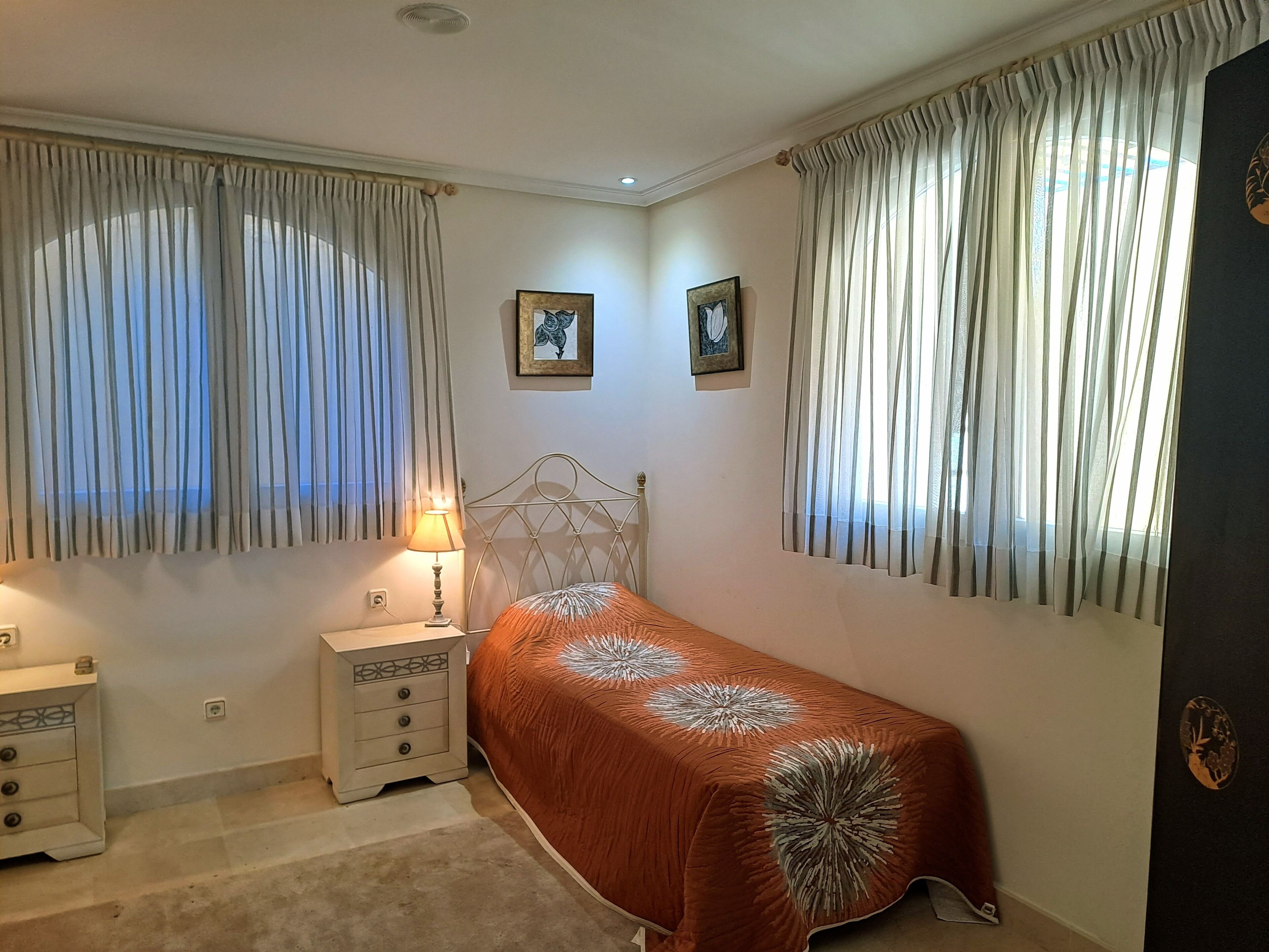 Un dormitorio con una cama individual, una mesita de noche con lámpara, dos cuadros en la pared y cortinas translúcidas en las ventanas.