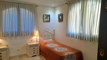Un dormitorio con una cama individual, una mesita de noche con lámpara, dos cuadros en la pared y cortinas translúcidas en las ventanas.