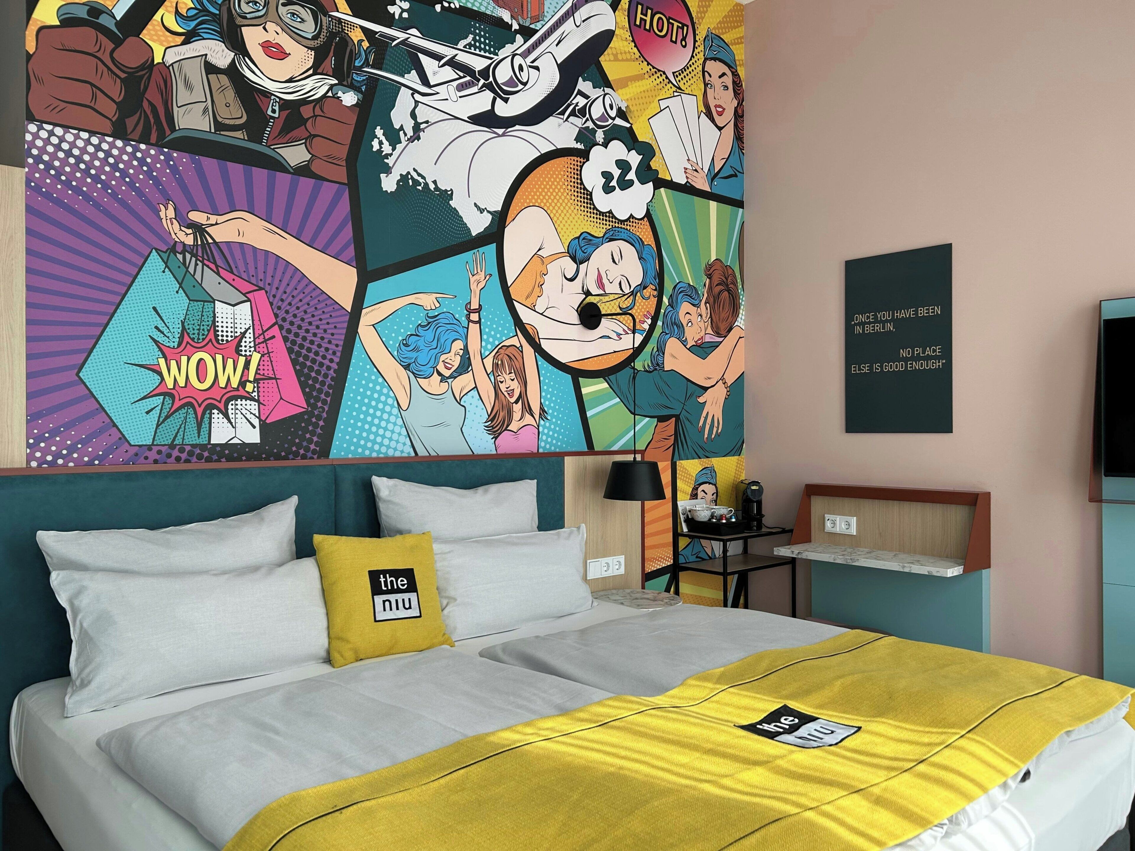 A hotel room with a bed, a wall mural, a coffee machine, and a quote on the wall.