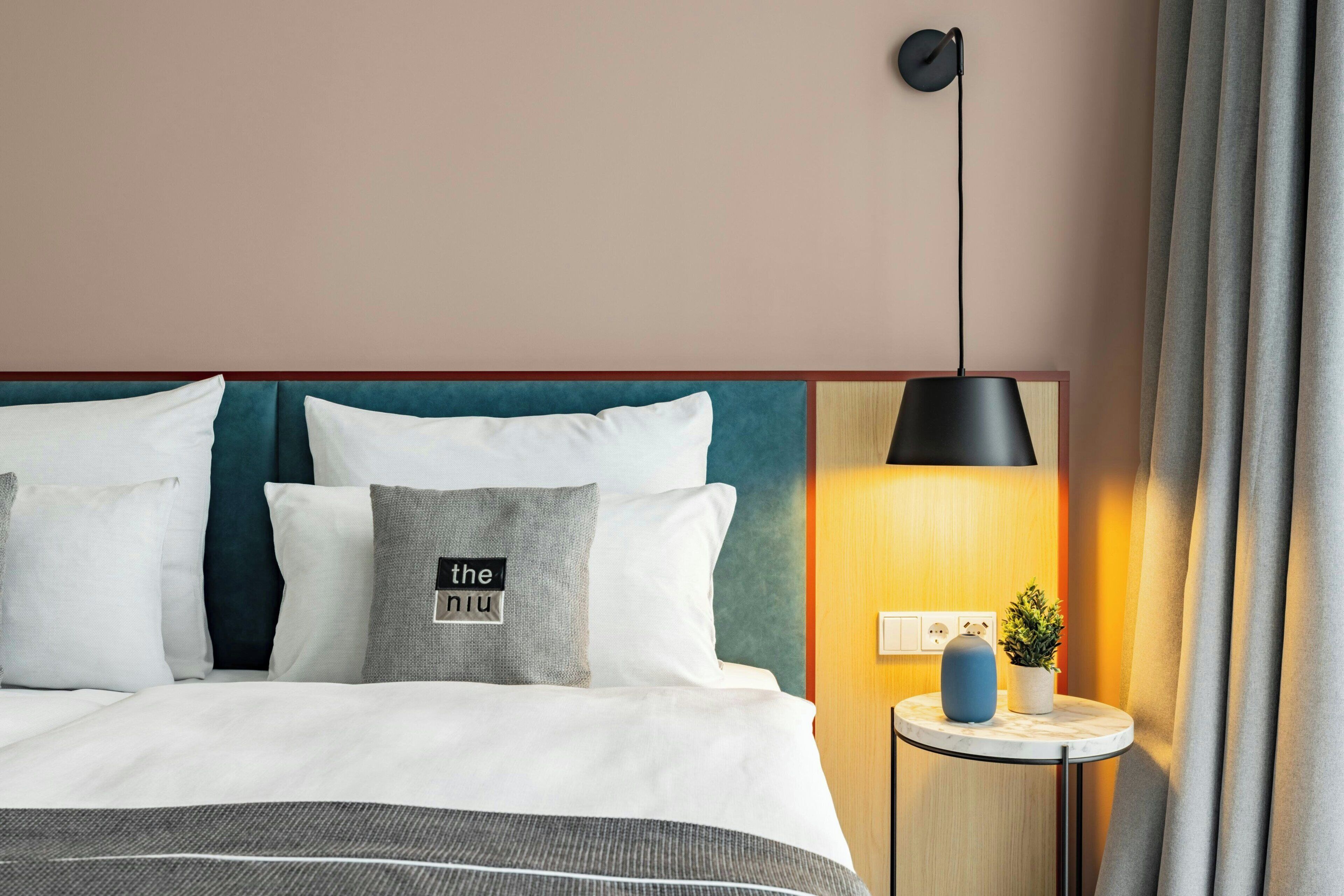 Una habitación de hotel con una cama, almohadas, una mesita de noche con una lámpara y una lámpara de pared.